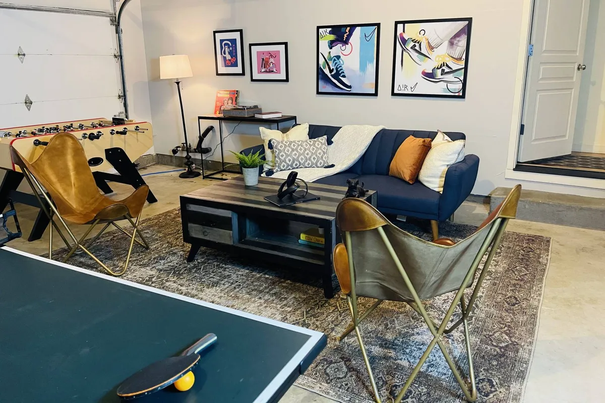 The photo shows a spacious game room with a sofa, wall art, a coffee table, and both a foosball and ping pong table. The layout emphasizes group gathering and casual play, with a comfortable, contemporary design.