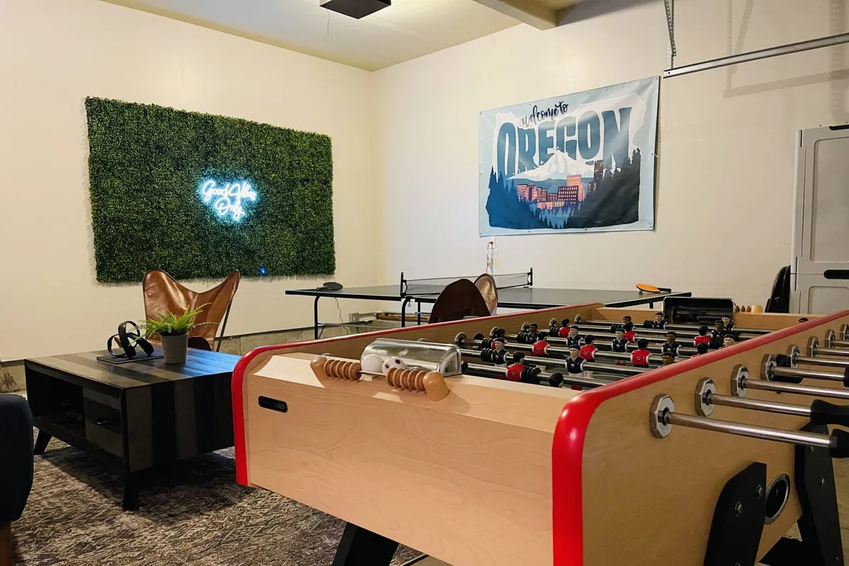 A lively interior scene with a foosball table, green wall featuring a neon sign, Oregon-themed artwork, and a comfortable seating area with chairs. The space balances work and play, offering room to relax and recharge.
