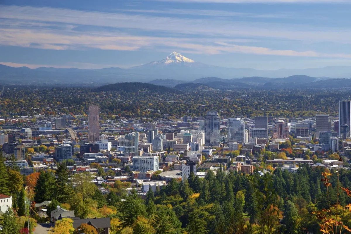 A panoramic view of the downtown Portland skyline with Mount Hood rising in the background, framed by lush trees in the foreground. The image captures the region's vibrant urban life and its scenic natural beauty, emphasizing accessibility and connection to local attractions.