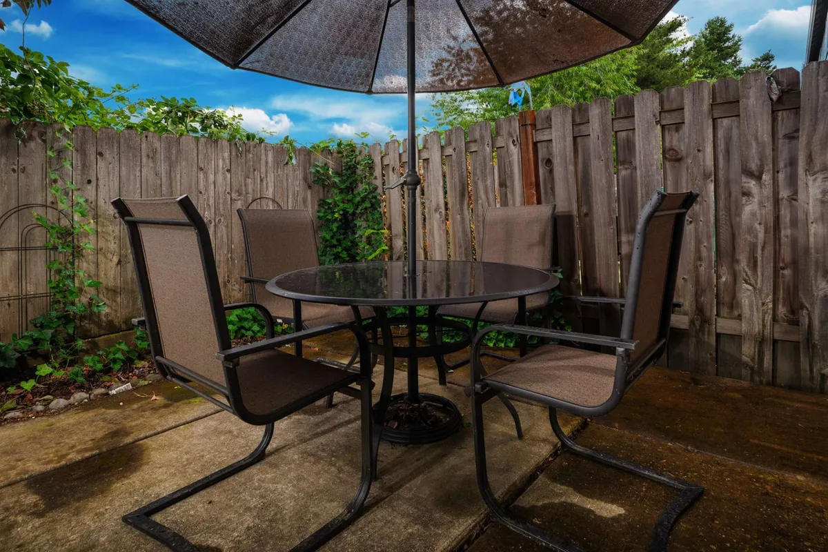 A sunny outdoor table set with an umbrella sits amid vibrant green foliage, bordered by a wooden fence, providing a cheerful and social space. The scene evokes options for outdoor dining and relaxation, perfect for unwinding after work or meeting up with colleagues or friends in a casual atmosphere.