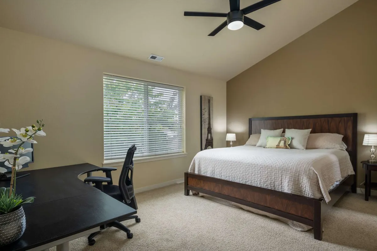A spacious master bedroom featuring a king-sized bed with crisp linens, a work desk and chair, a large window with blinds letting in natural light, and nightstands with lamps for evening comfort.