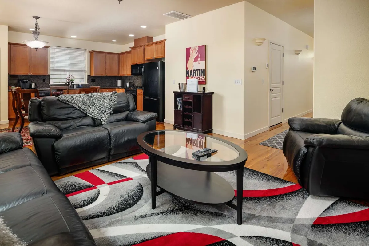 A modern, open-plan living room featuring black leather sofas, a glass coffee table, and an art deco poster. The kitchen area is visible in the background, showcasing the flow between living and dining spaces. The setup suggests a space perfect for socializing, hosting, and casual entertainment, with ample room for group activities.
