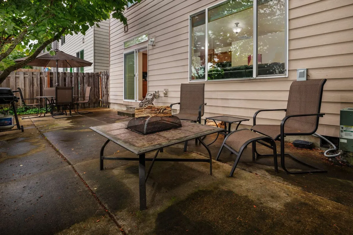 An inviting exterior patio scene highlights a table with outdoor chairs, lush tree coverage, a barbecue grill, and serene greenery—all enclosed within a fence for privacy. The setting is staged for outdoor dining, social gatherings, or a peaceful coffee break.