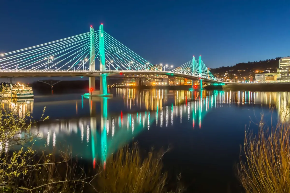 A tranquil evening scene featuring an illuminated bridge over calm water, with reflections of city lights and a hint of nearby activity. The view suggests accessibility to vibrant outdoor spaces and local entertainment options.
