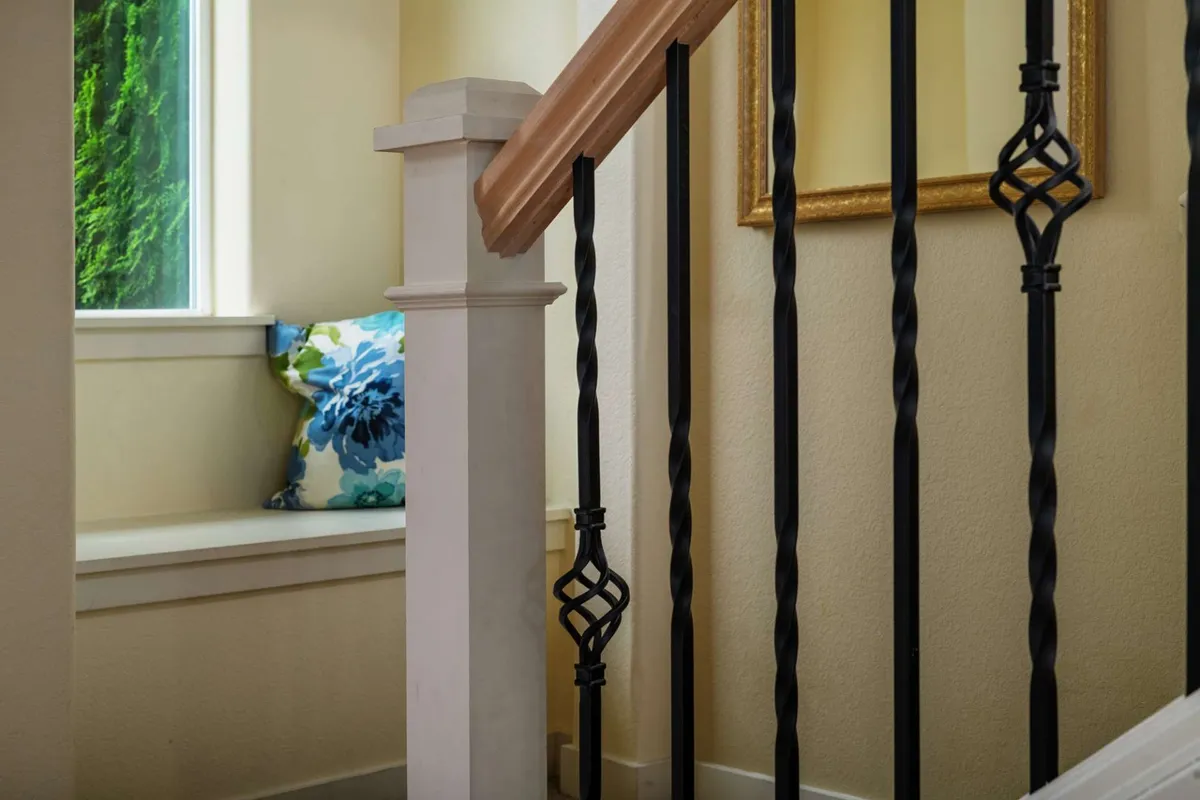 A bright, welcoming window seat with a decorative pillow, adjacent to a staircase with elegant railing and framed artwork. The scene suggests comfortable conversation, reading, or a cozy rendezvous before heading out for the day.