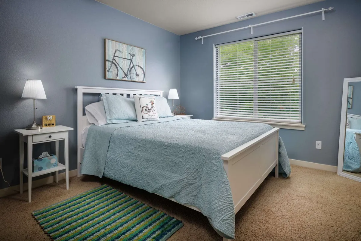 A serene bedroom with a bed dressed in a blue quilt, bicycle-themed wall art, a window with blinds for adjustable light, and white bedside tables for added comfort.